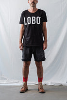 Man wearing a black crew neck t-shirt with Lobo logo, black denim shorts, high red socks, and brown leather boots.