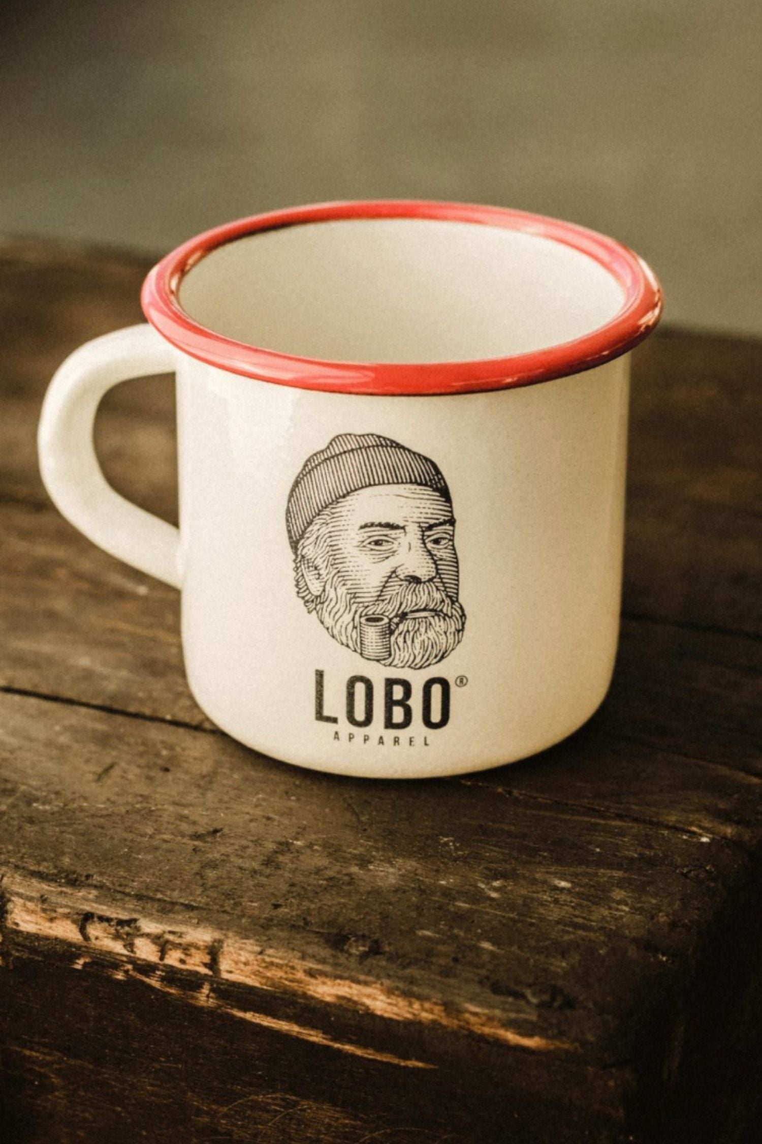 White enamel mug from LOBO Apparel with a red rim, featuring the illustration of a man wearing a beanie and smoking a pipe, set on an antique wooden box.