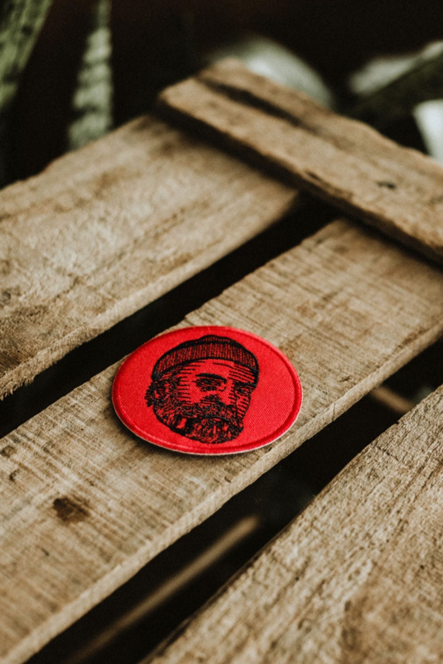 Round red Lobo Face embroidered patch with a bearded man design placed on rustic wooden slats.