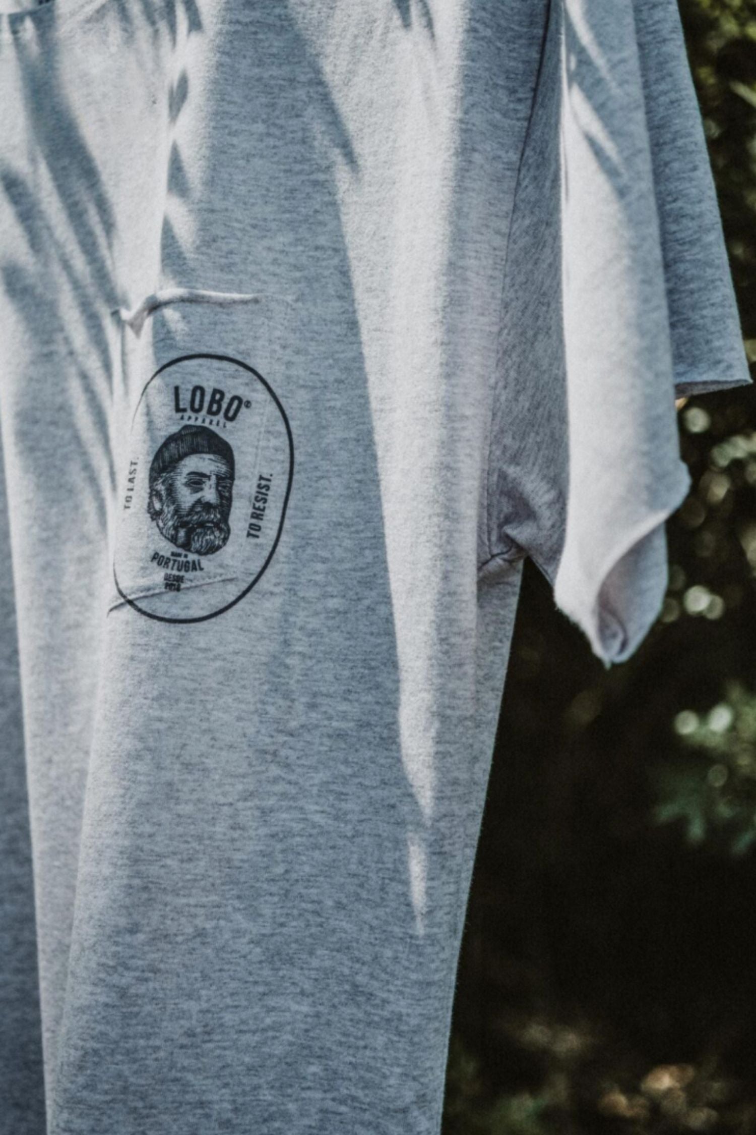 Close-up detail of the grey t-shirt's chest pocket, showing the screen-printed Lobo emblem with a sailor illustration and the phrases "To Last" and "To Resist".