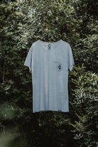 Unisex Lobo grey t-shirt hanging on a hanger outdoors, featuring the chest pocket logo against a green foliage background.