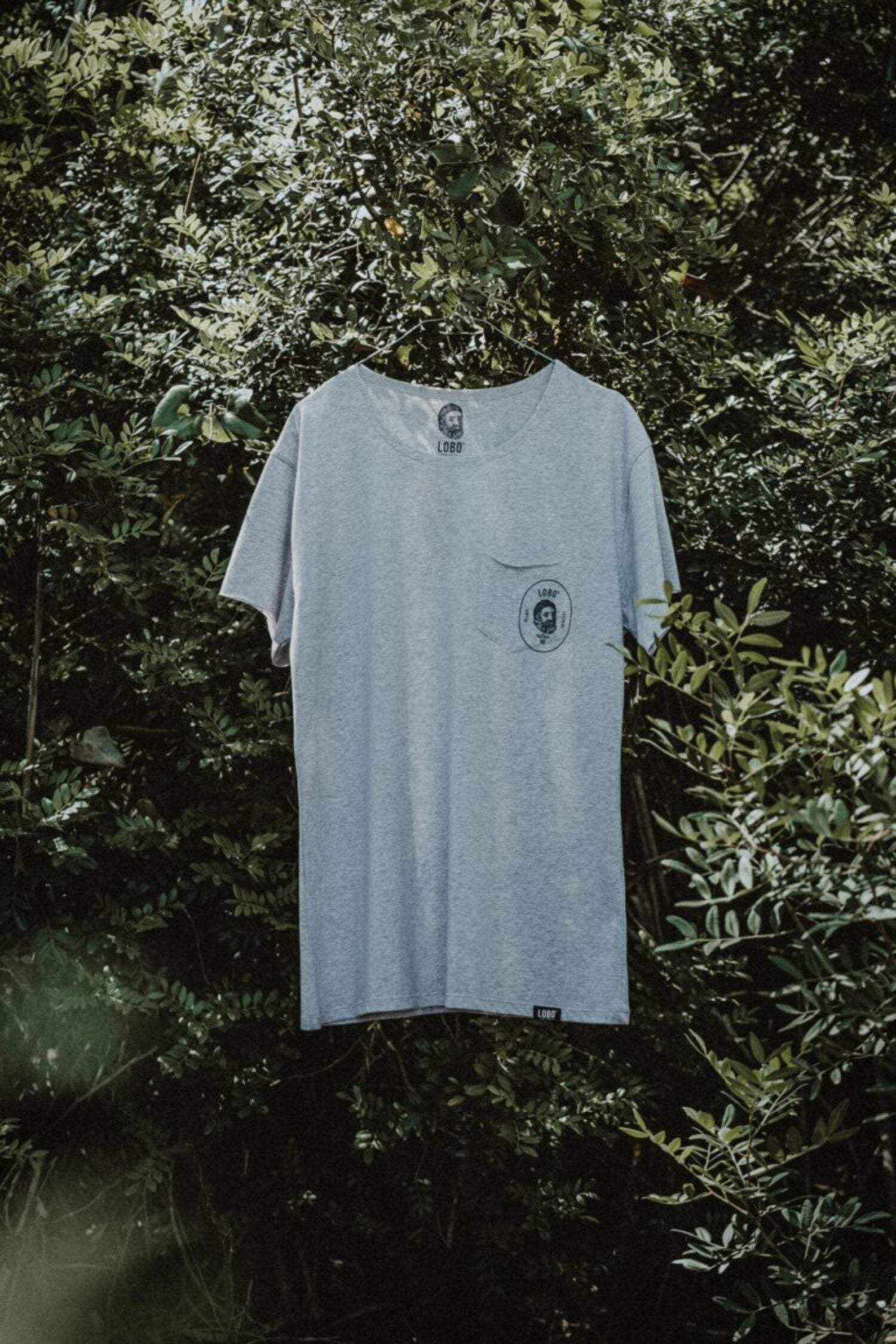 Unisex Lobo grey t-shirt hanging on a hanger outdoors, featuring the chest pocket logo against a green foliage background.