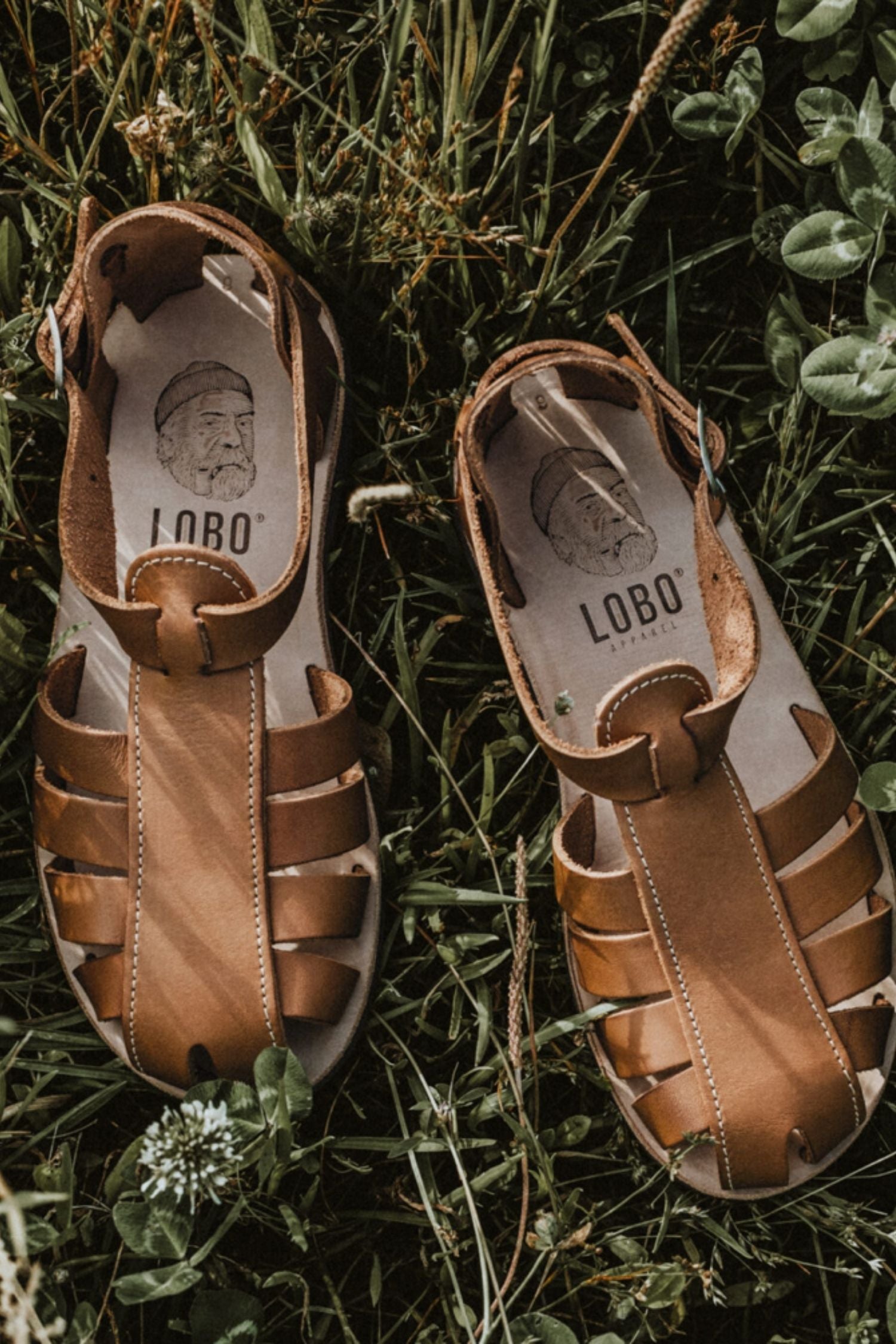 Lobo brown leather sandals with rubber soles on green grass, featuring the leather insole with the brand's logo and illustration.
