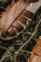Close-up of Lobo brown leather sandals on grass, highlighting the detailed white stitching and the premium leather finish.