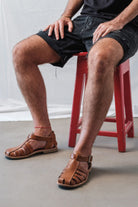 Man sitting on a red stool wearing brown Lobo Leather Sandals, demonstrating the comfortable fit and minimalist leather strap design.