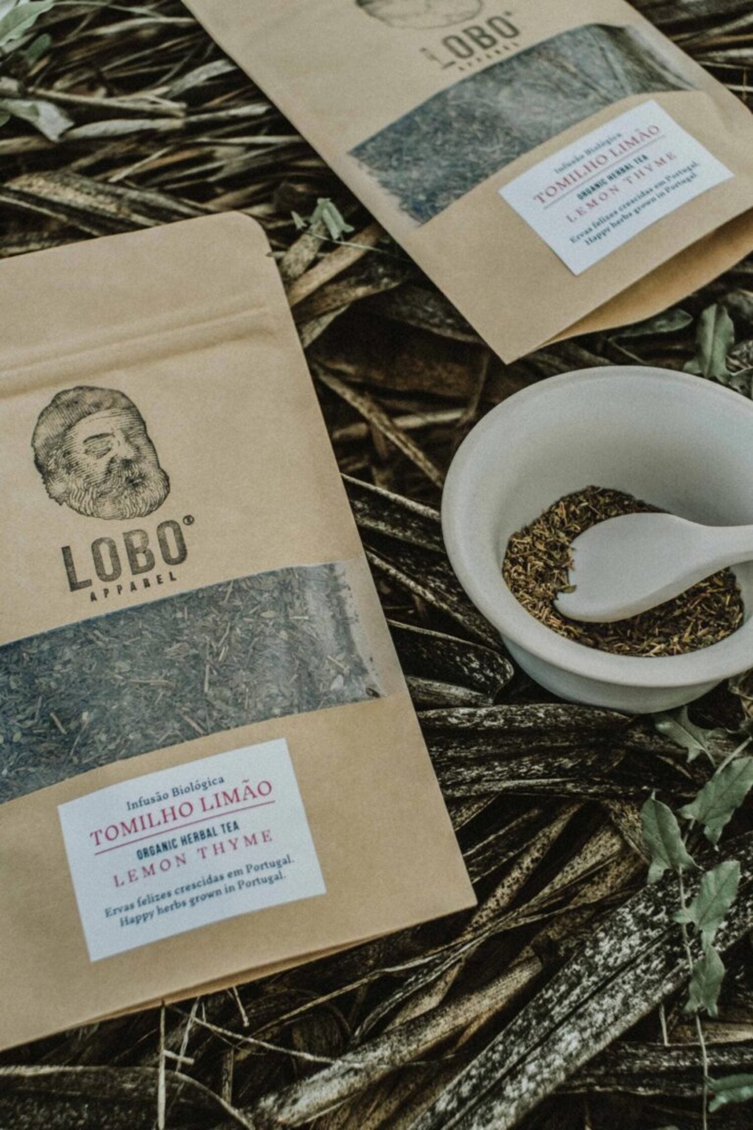 Two packages of Lobo Organic Lemon Thyme herbal tea next to a bowl of dried herbs, arranged on a natural ground of leaves and twigs.