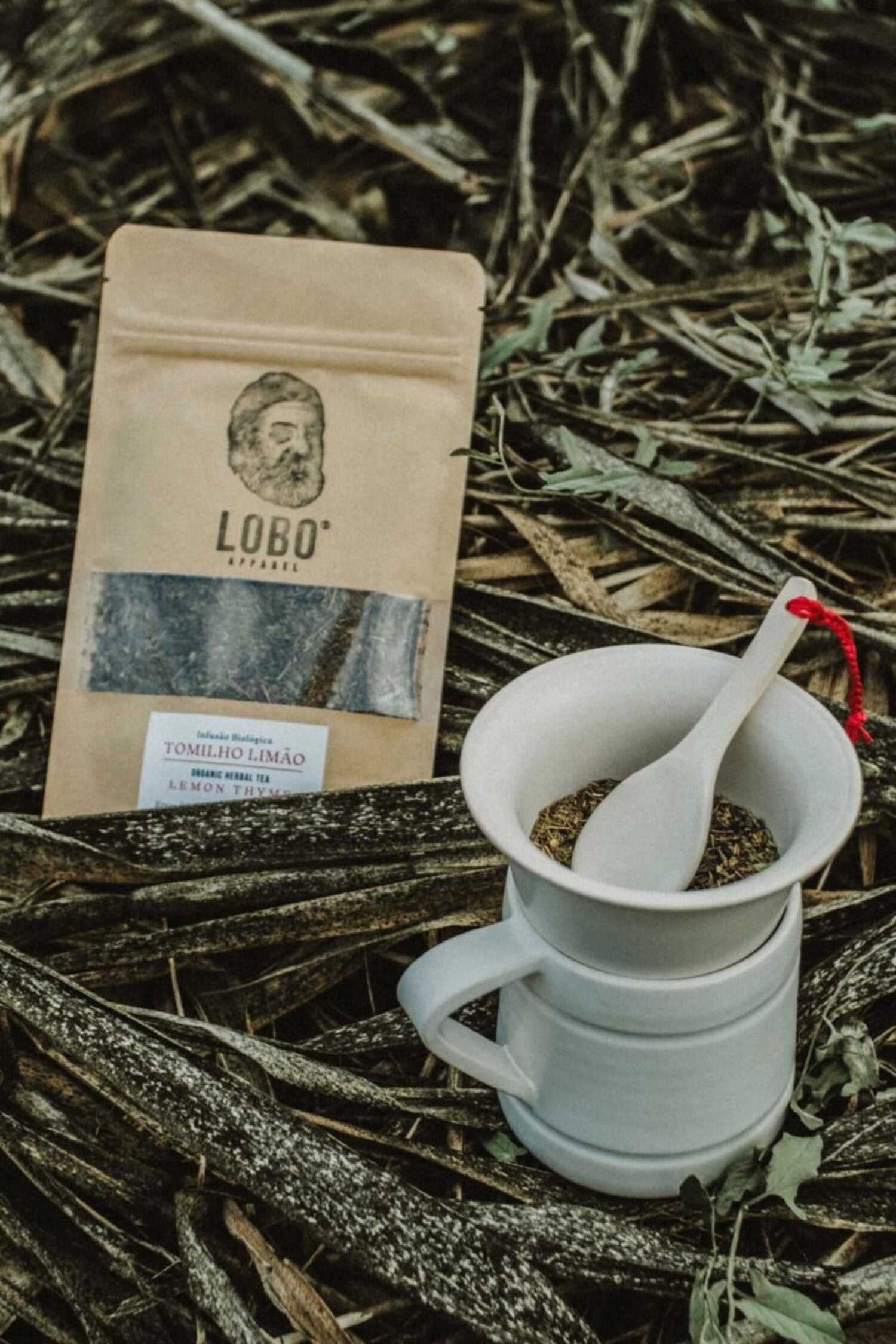 Top-down view of a Lobo Lemon Thyme tea pouch next to a ceramic bowl with herbs and a white spoon, placed on dry straw in a natural outdoor setting.