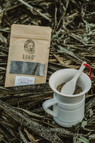 Top-down view of a Lobo Lemon Thyme tea pouch next to a ceramic bowl with herbs and a white spoon, placed on dry straw in a natural outdoor setting.
