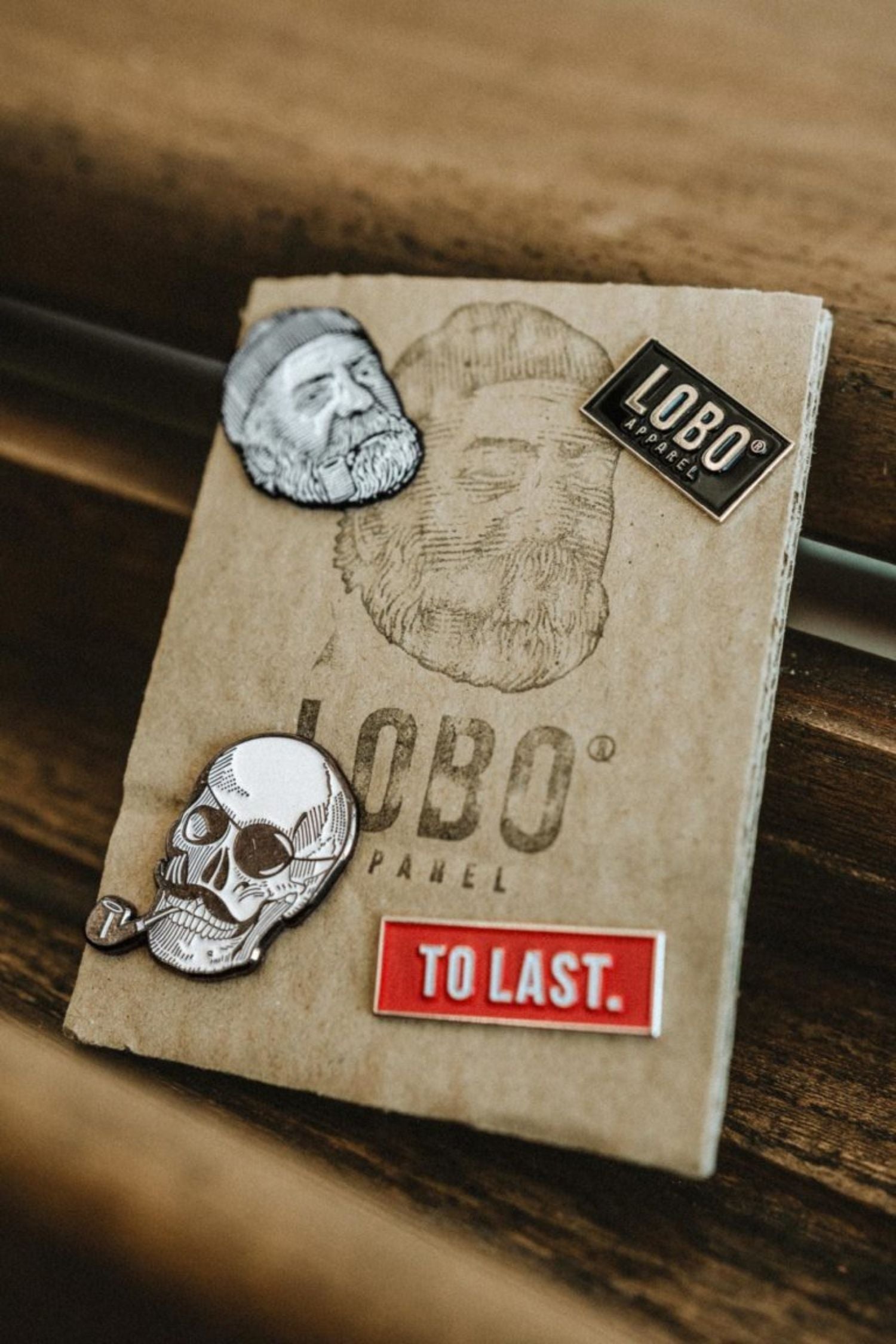 Black Lobo Logo metal pin on Lobo Apparel kraft packaging, alongside the red To Last pin and skull pin from the collection. 
