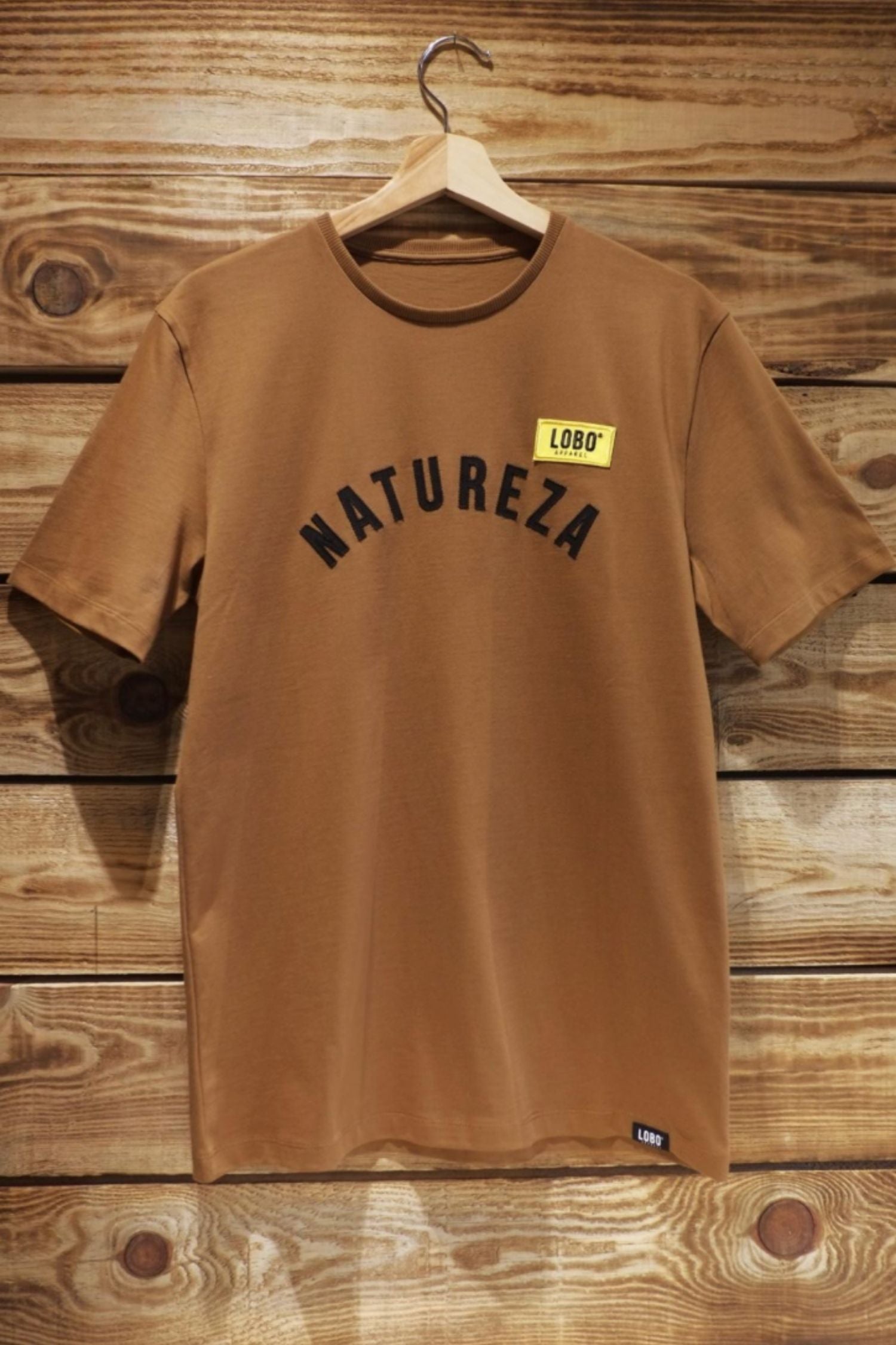 Unisex Lobo "Natureza" t-shirt in brown, hanging on a wooden hanger against a rustic wooden wall.