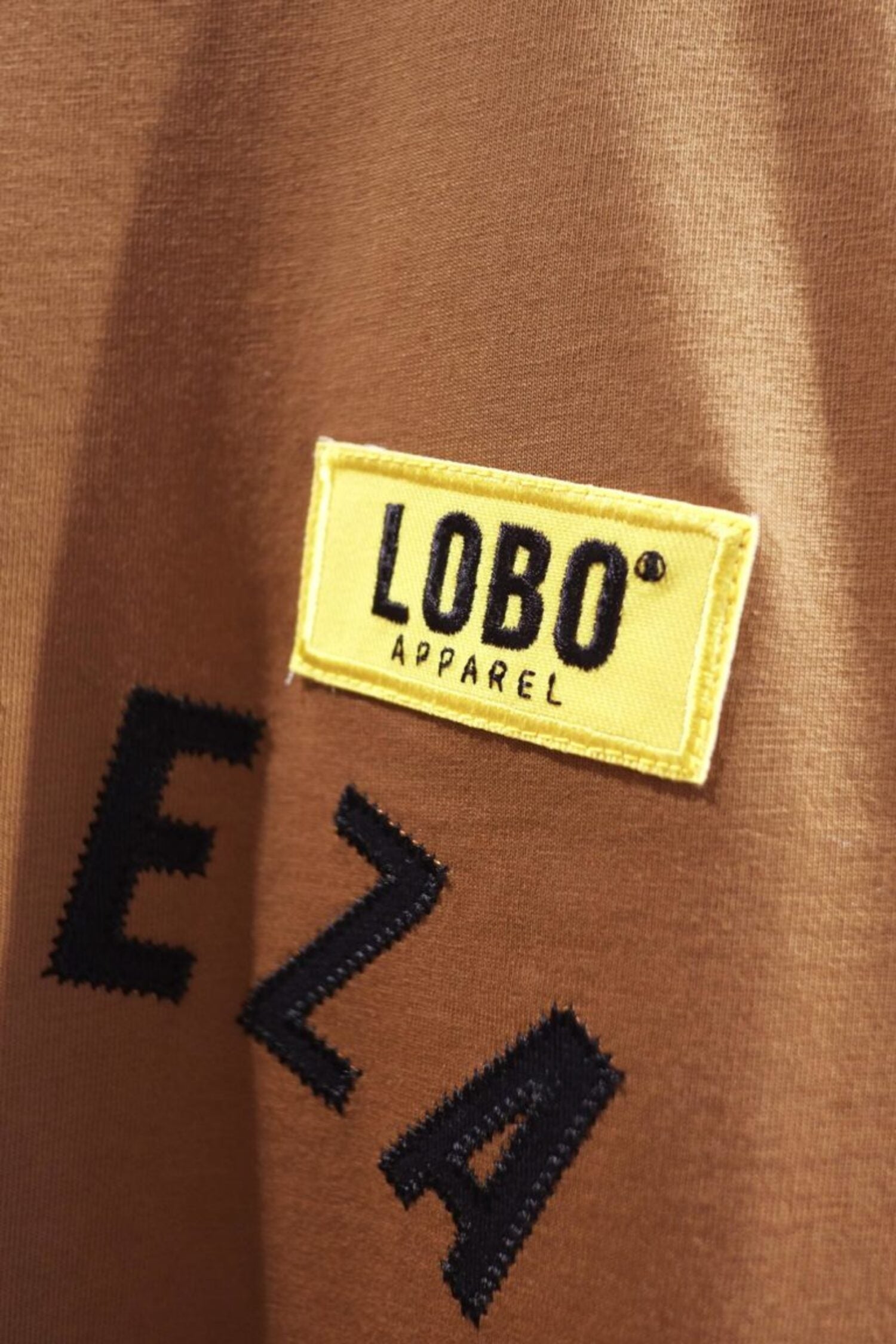 Close-up of the yellow rectangular patch with the black "LOBO Apparel" logo sewn onto the chest of the brown t-shirt.