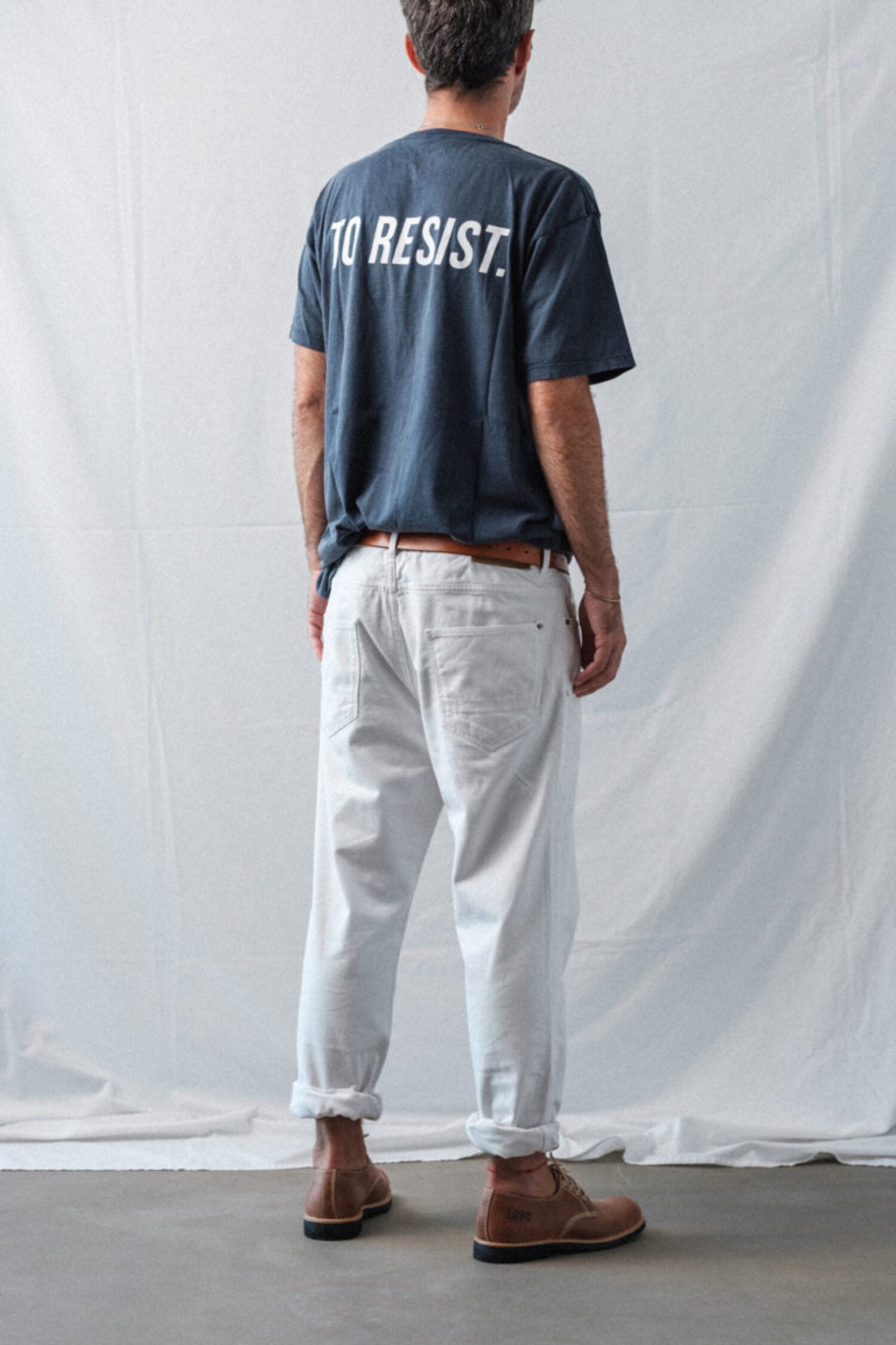 Back view of a man wearing Lobo Raw shoes, white trousers, and a blue t-shirt with "To Resist" text.