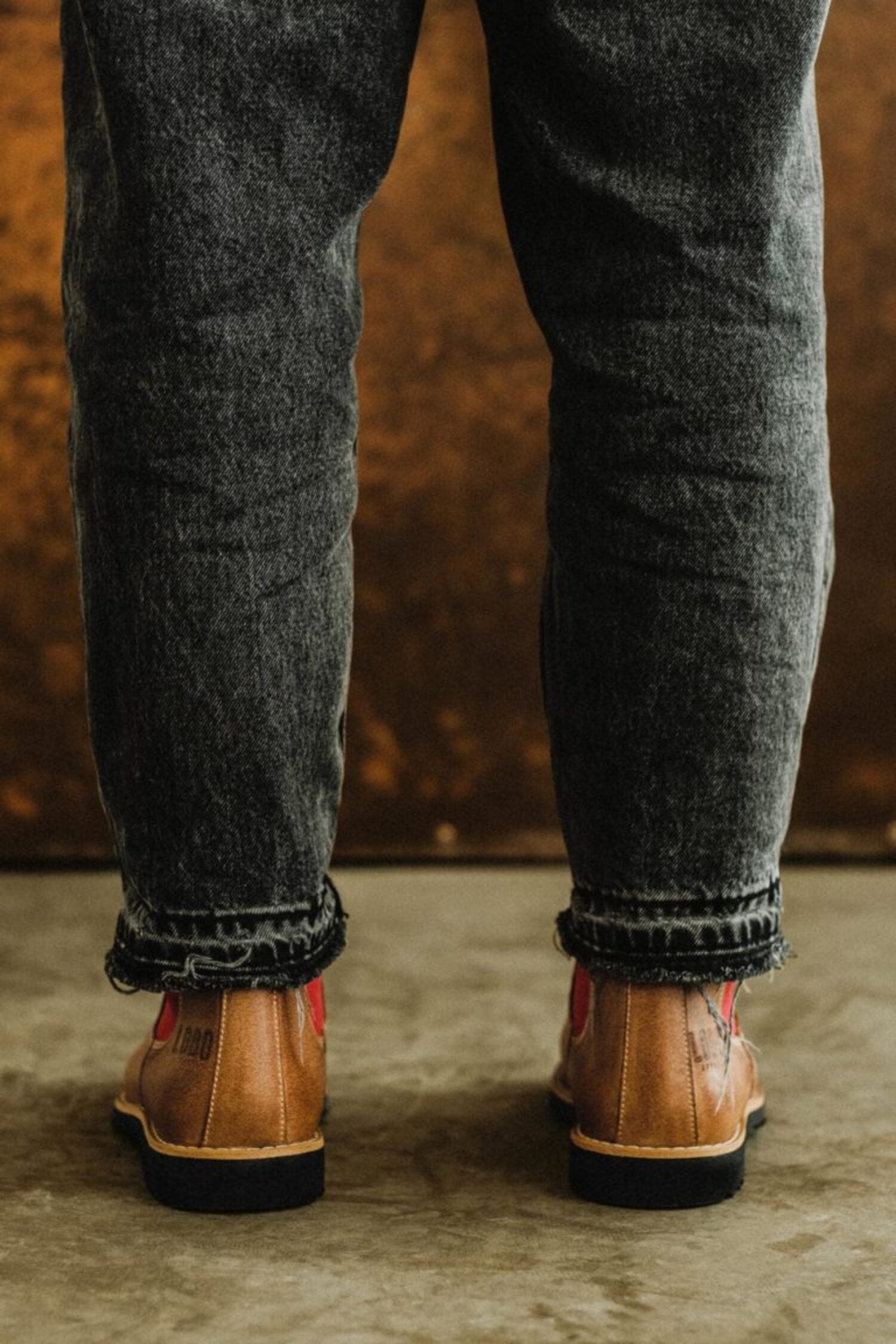 Rear view of a person wearing Lobo tan leather boots with dark wash jeans, showing red side panels.