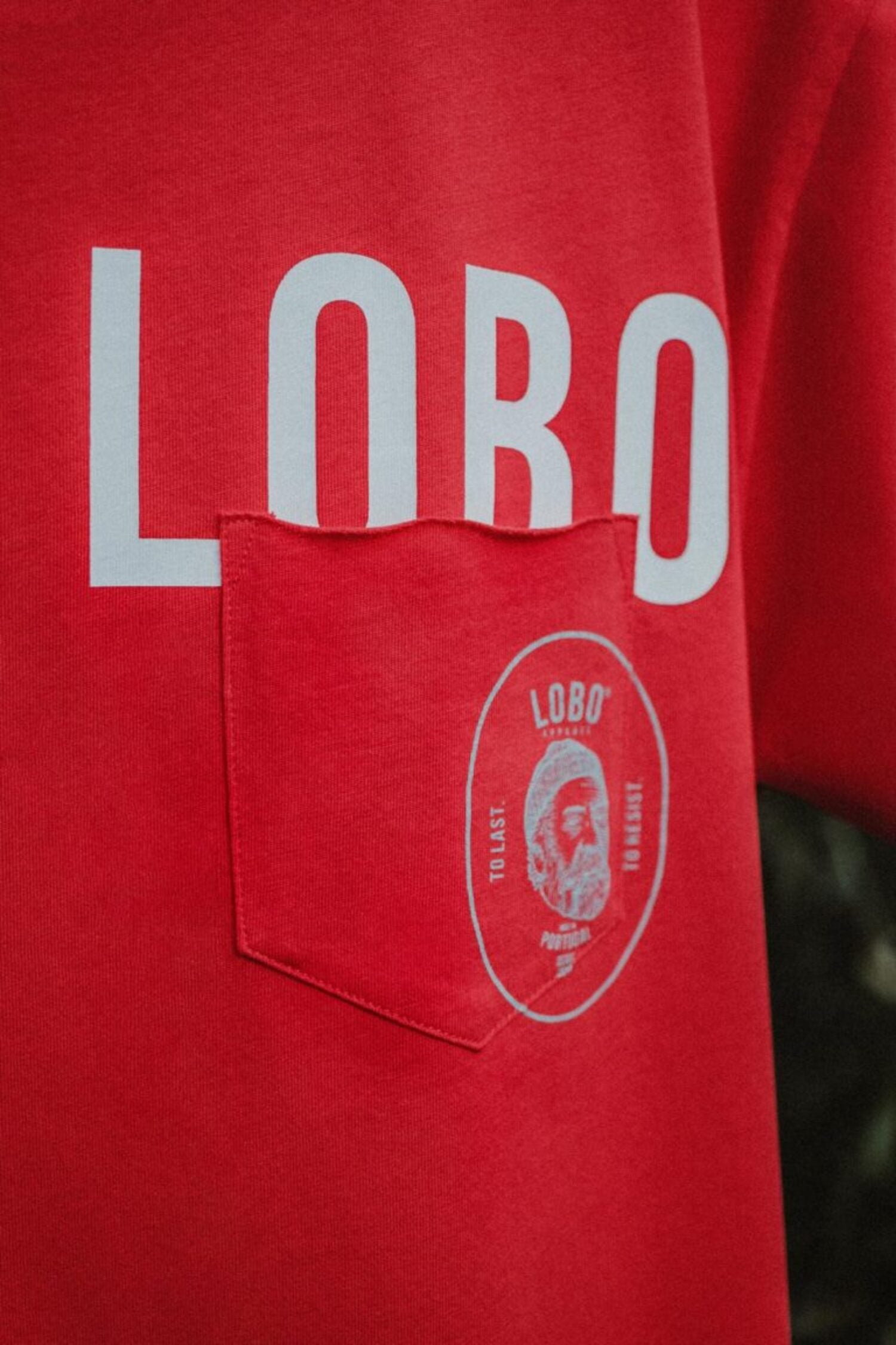 Detail of a red pocket t-shirt showcasing the "LOBO" brand name in bold letters and a circular seal featuring the man with a beanie and pipe.