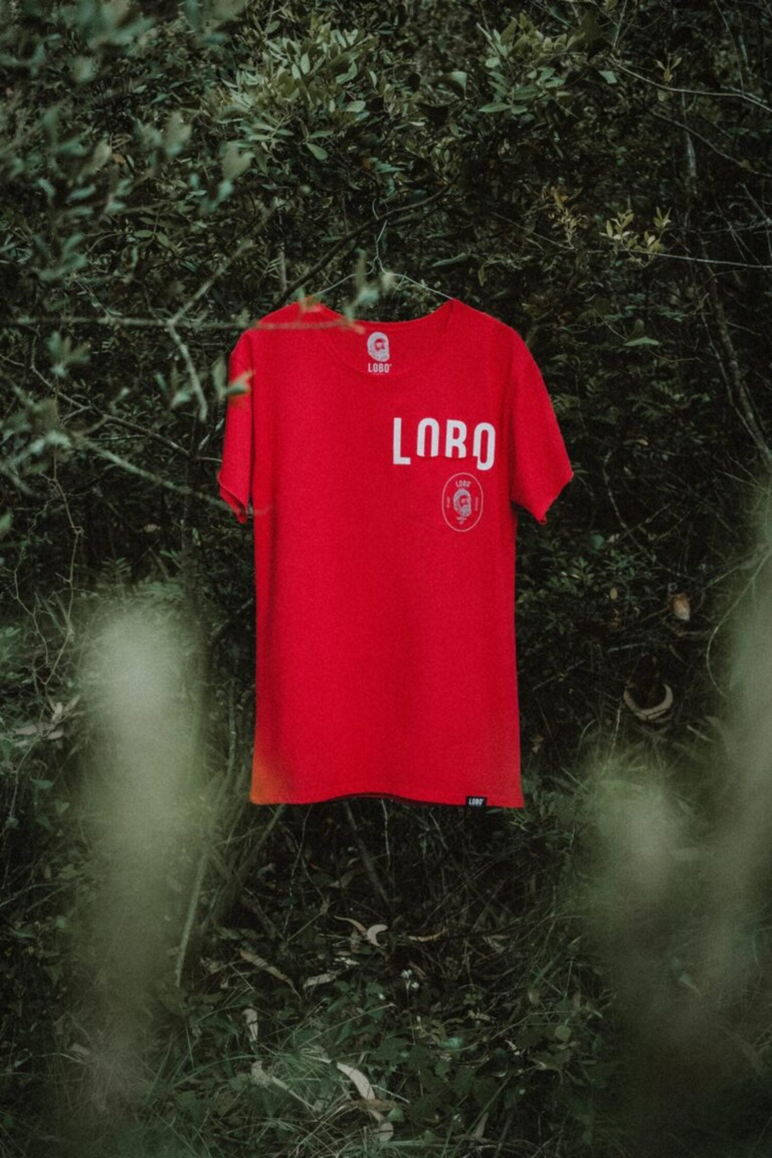 Lobo red premium cotton t-shirt hanging on a tree branch, showing the "LOBO" screen print and front pocket detail.