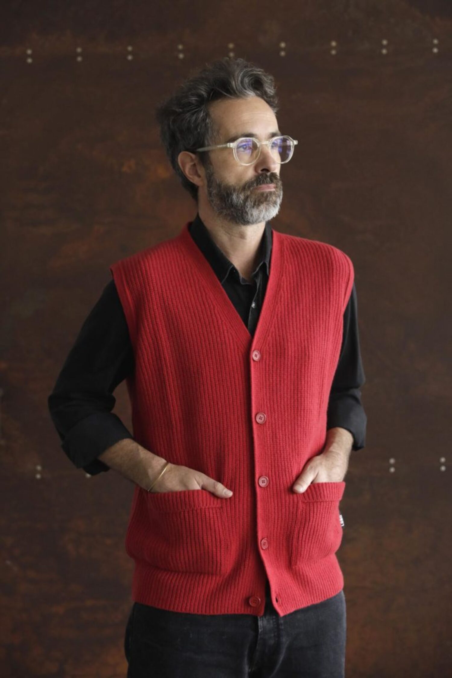 Bearded man with glasses wearing the Lobo Red Vest, a unisex red wool vest, over a black shirt against a rustic background.