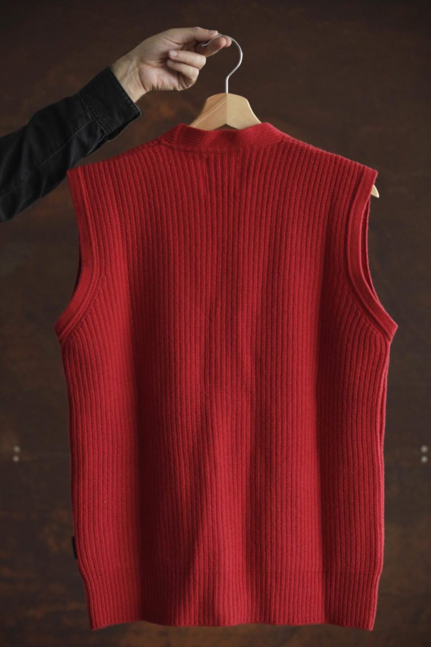Back view of the red wool Lobo Red Vest hanging on a wooden hanger, highlighting the continuous ribbed knit and the unisex cut.