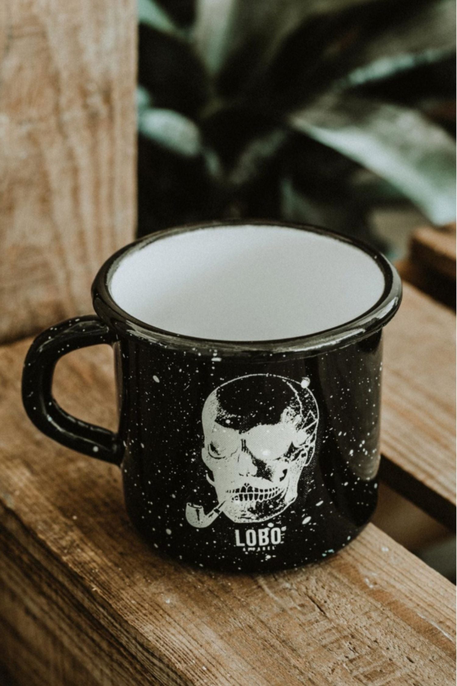 Lobo Skull black enamel mug featuring a skull smoking a pipe illustration, resting on rustic wood.