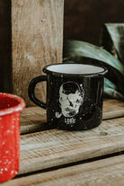 Black Lobo Skull Enamel mug with skull illustration on a wooden surface, featuring a partial red mug and plant in the background.