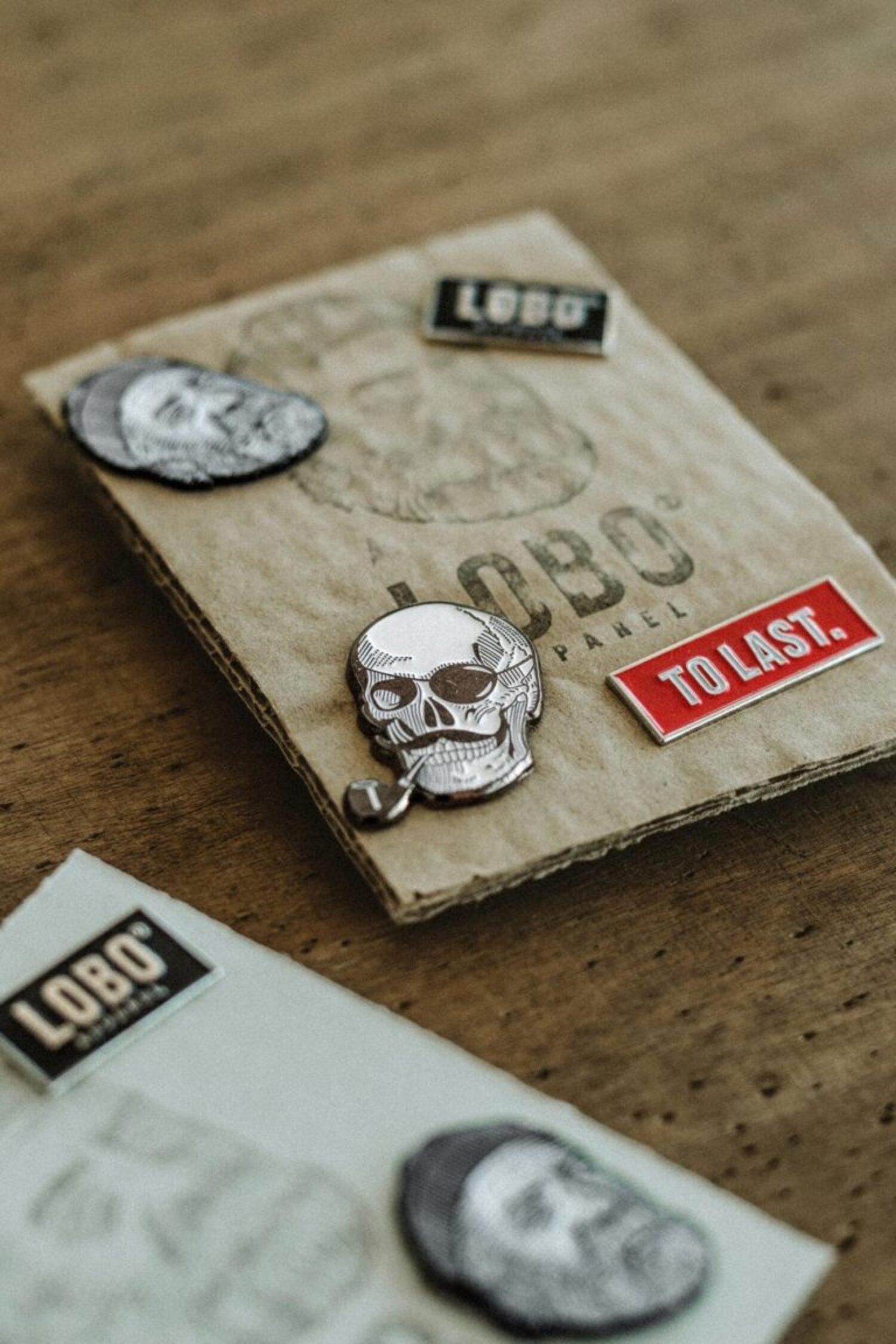 Collection of Lobo Apparel metal pins, including the Lobo Skull Pin and red "To Last" pin, displayed on sustainable cardboard packaging.