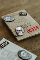 Collection of Lobo Apparel metal pins, including the Lobo Skull Pin and red "To Last" pin, displayed on sustainable cardboard packaging.