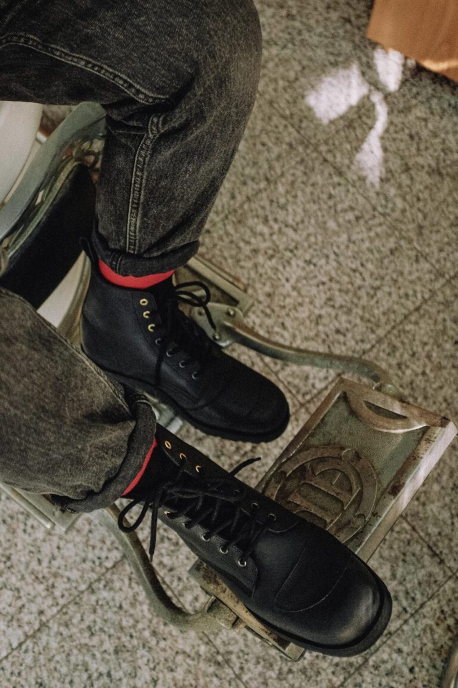 High-angle view of the black leather Lobo Tigre Boots 3rd Edition, worn with red socks and grey denim jeans, highlighting the golden eyelets and the motorcycle shifter protector.