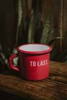 "To Last" red enamel mug on a rustic wooden table with a plant in the background.