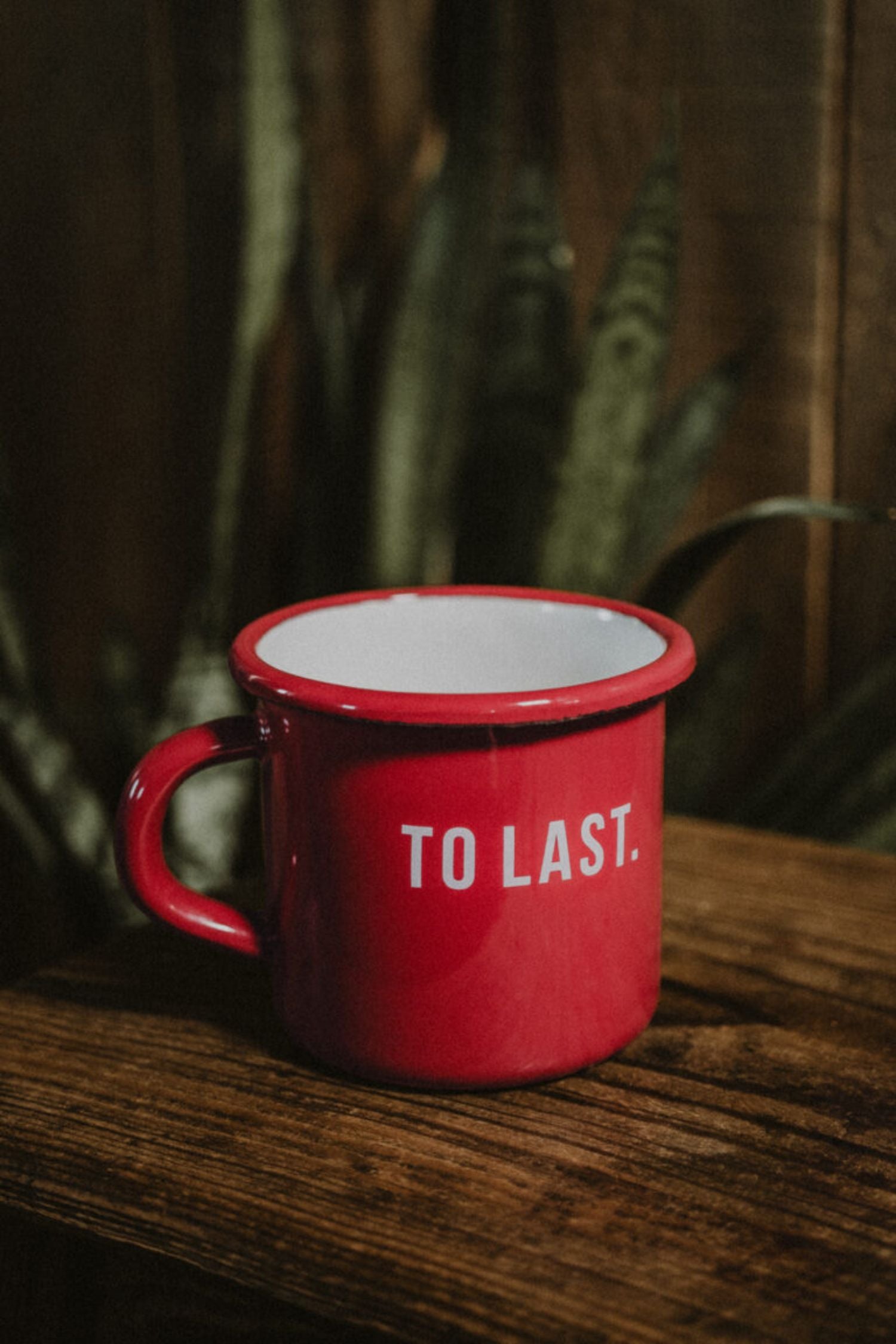 "To Last" red enamel mug on a rustic wooden table with a plant in the background.