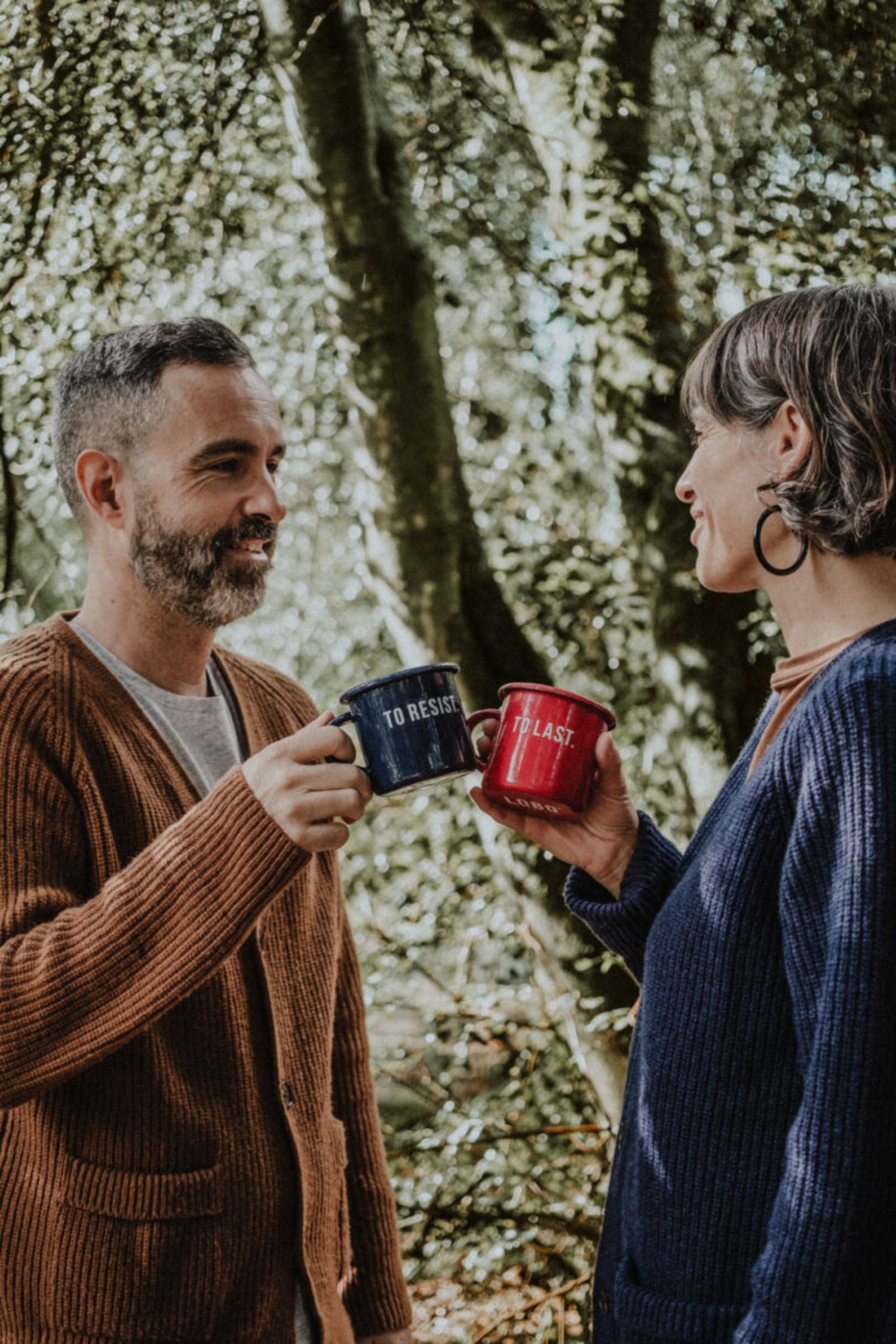 Couple toasting with Lobo "To Resist" blue and "To Last" red enamel mugs in a forest setting.