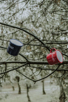 Blue and red Lobo enamel mugs hanging from tree branches in a natural outdoor setting.