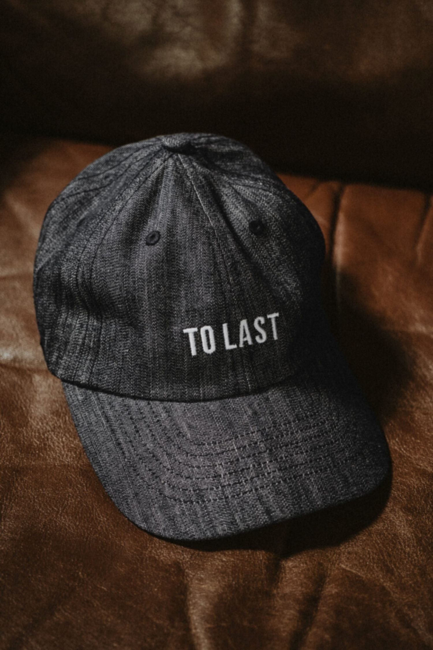 Lobo Apparel grey denim cap with "To Last" embroidered in white, resting on a brown leather surface.