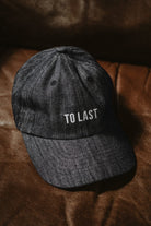 Lobo Apparel grey denim cap with "To Last" embroidered in white, resting on a brown leather surface.