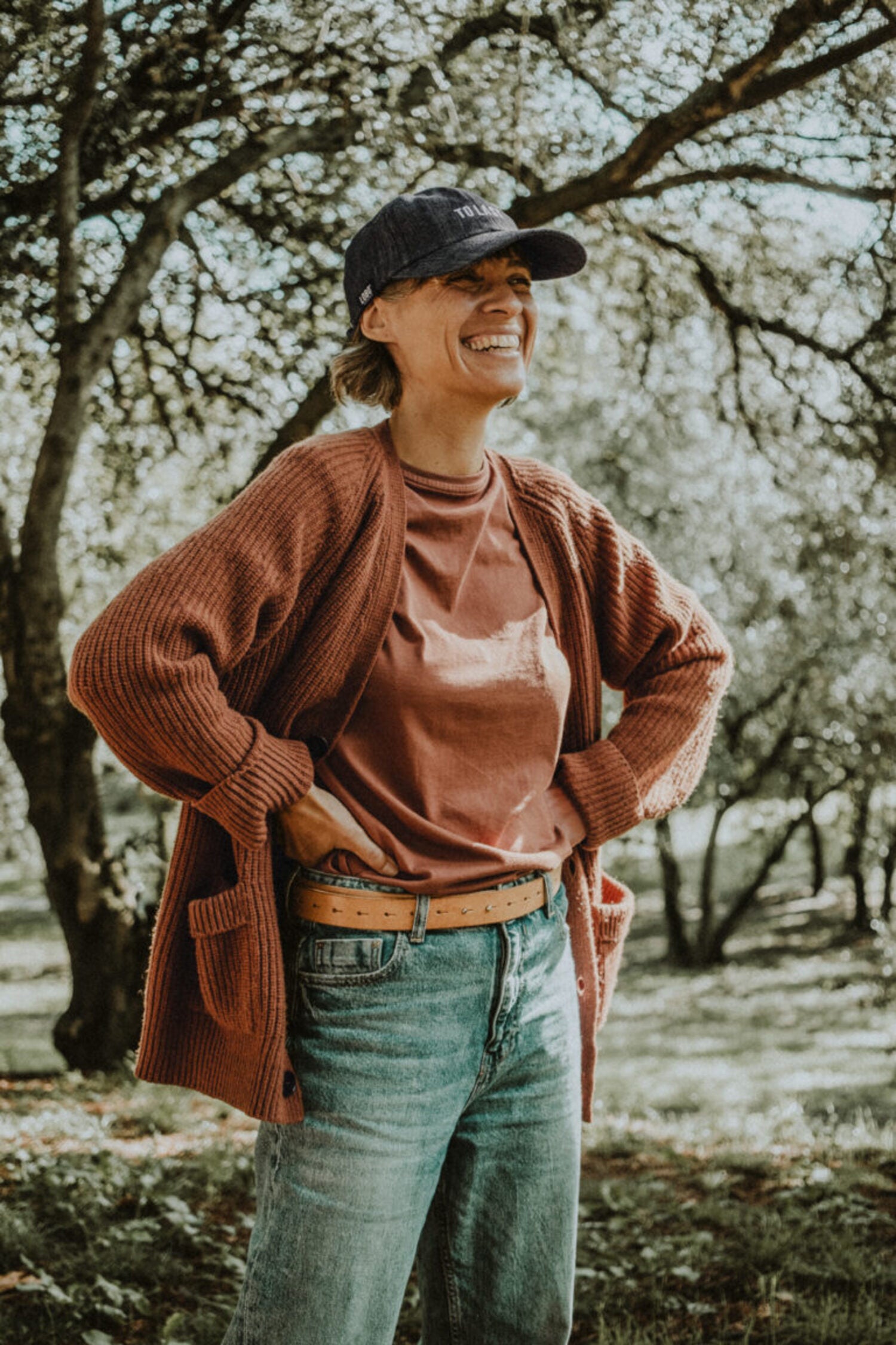 Smiling woman outdoors wearing the Lobo "To Last" grey denim cap, paired with a rust-colored cardigan.