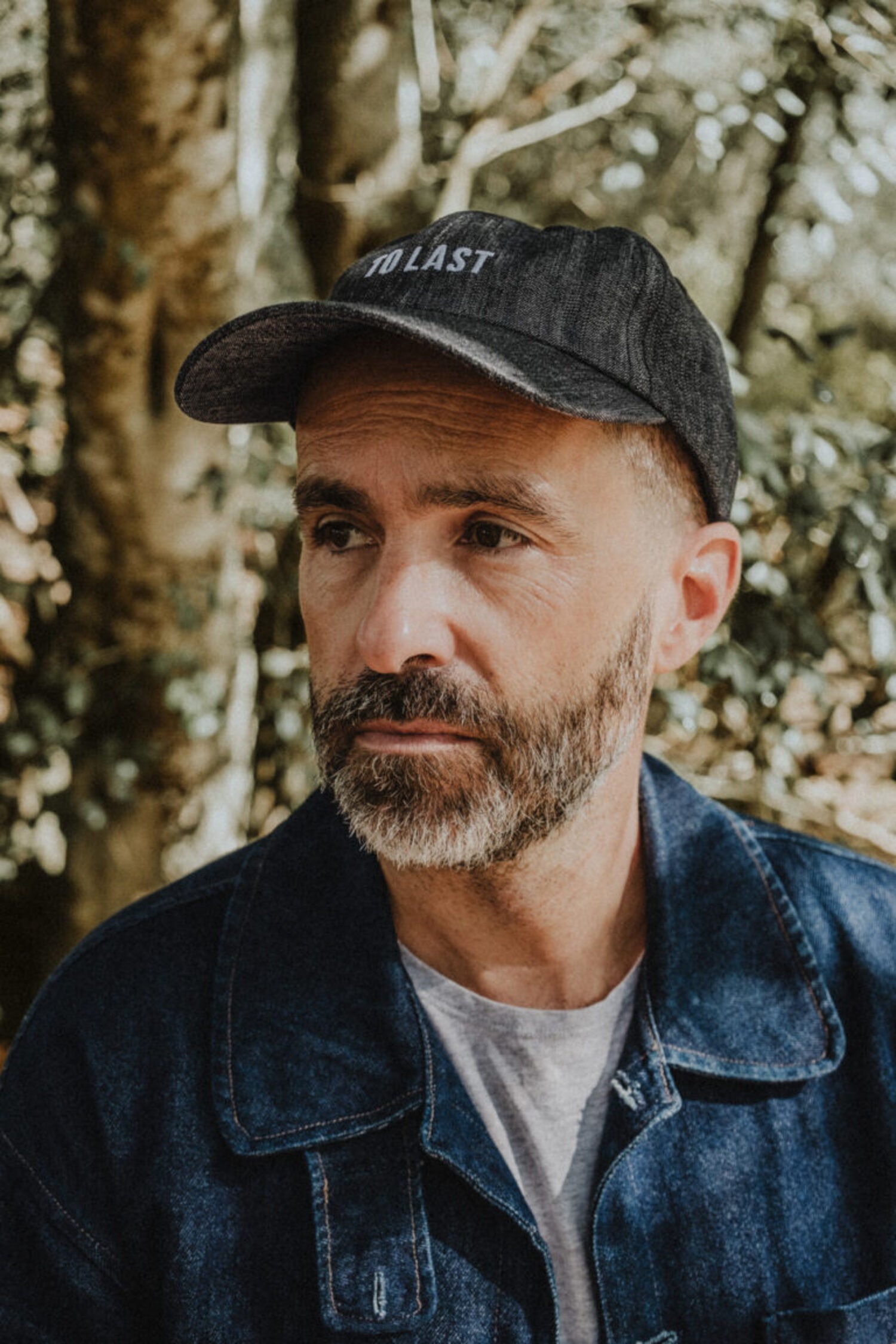Bearded man wearing the "To Last" grey denim cap and a blue denim jacket in a natural setting with trees in the background.