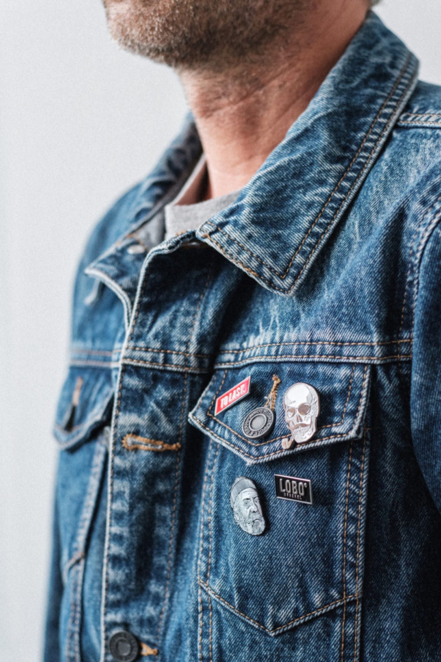 Detail of a denim jacket with several metal pins, including the red Lobo To Last pin and the Lobo Apparel pin.