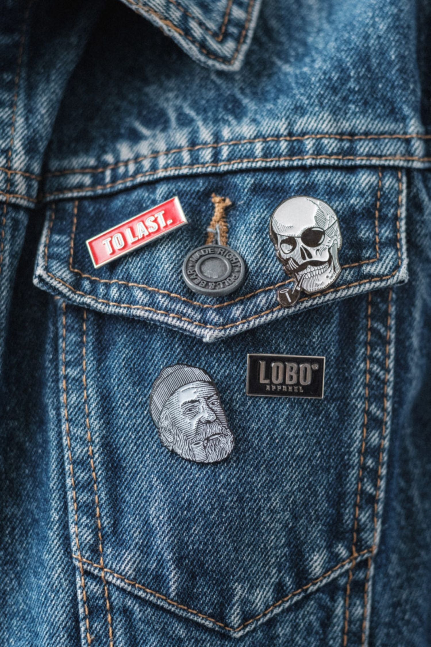 Close-up of a denim jacket pocket with four Lobo Apparel metal pins, including the red To Last pin, skull pin, round pin and face illustration pin.