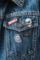 Close-up of a denim jacket pocket with four Lobo Apparel metal pins, including the red To Last pin, skull pin, round pin and face illustration pin.