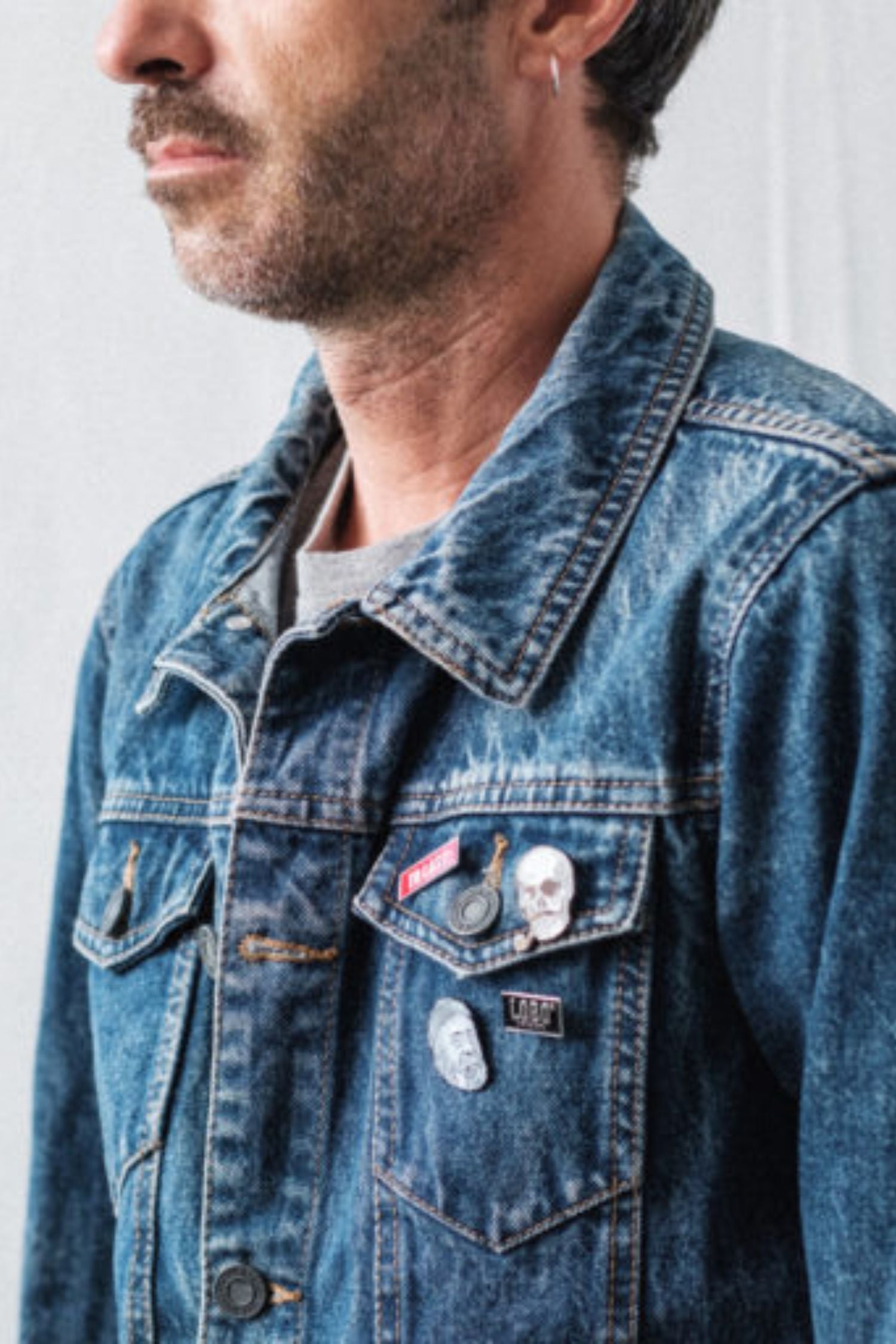 Man wearing a denim jacket with a set of Lobo Apparel metal pins on the front pocket, including the red To Last pin.