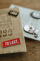 Red Lobo To Last metal pin resting on a Lobo Apparel kraft packaging, with other collection pins visible in the background.