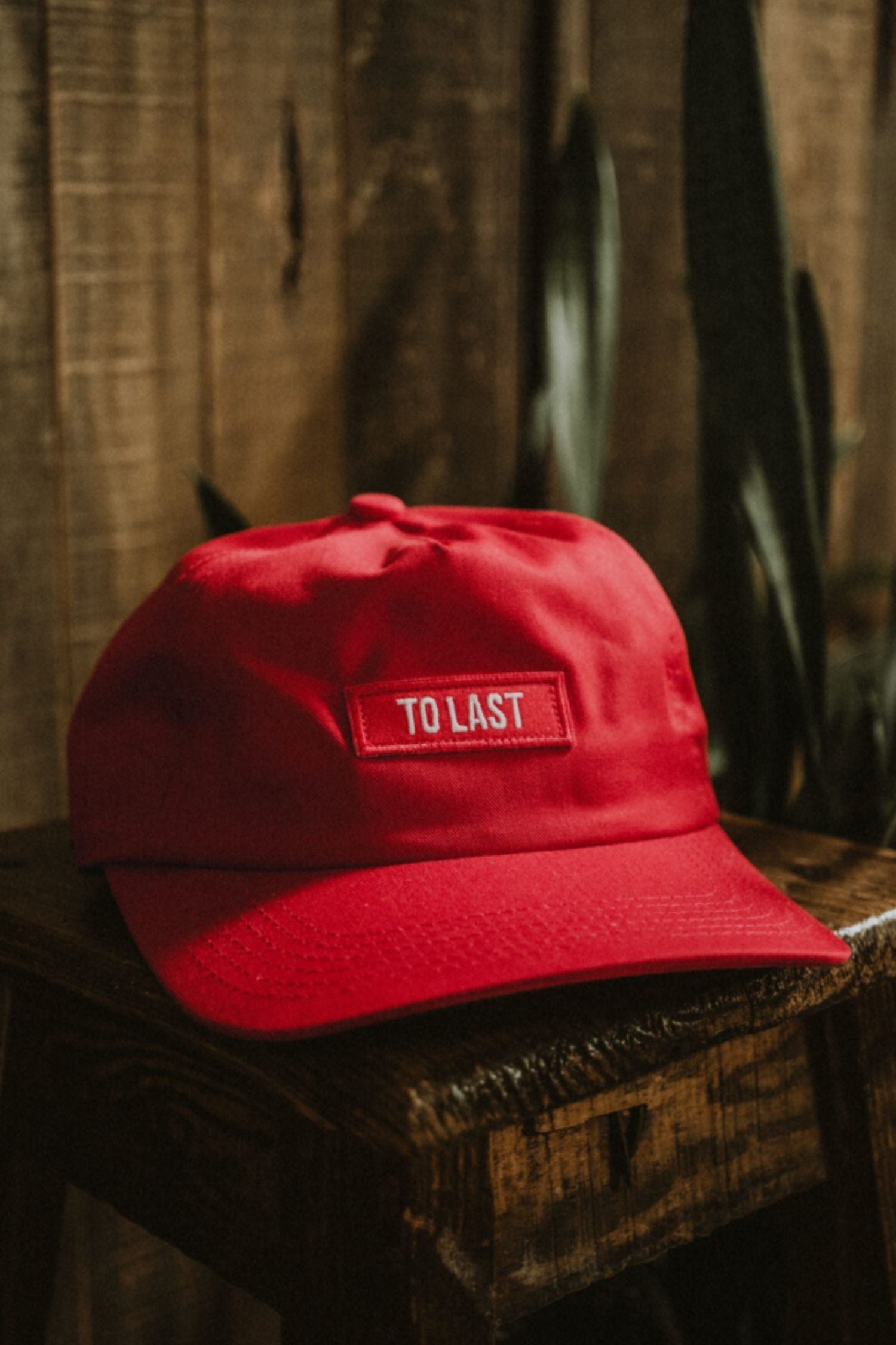 Red premium LOBO Apparel cap with an embroidered "TO LAST" patch on the front, set on a rustic wooden surface.