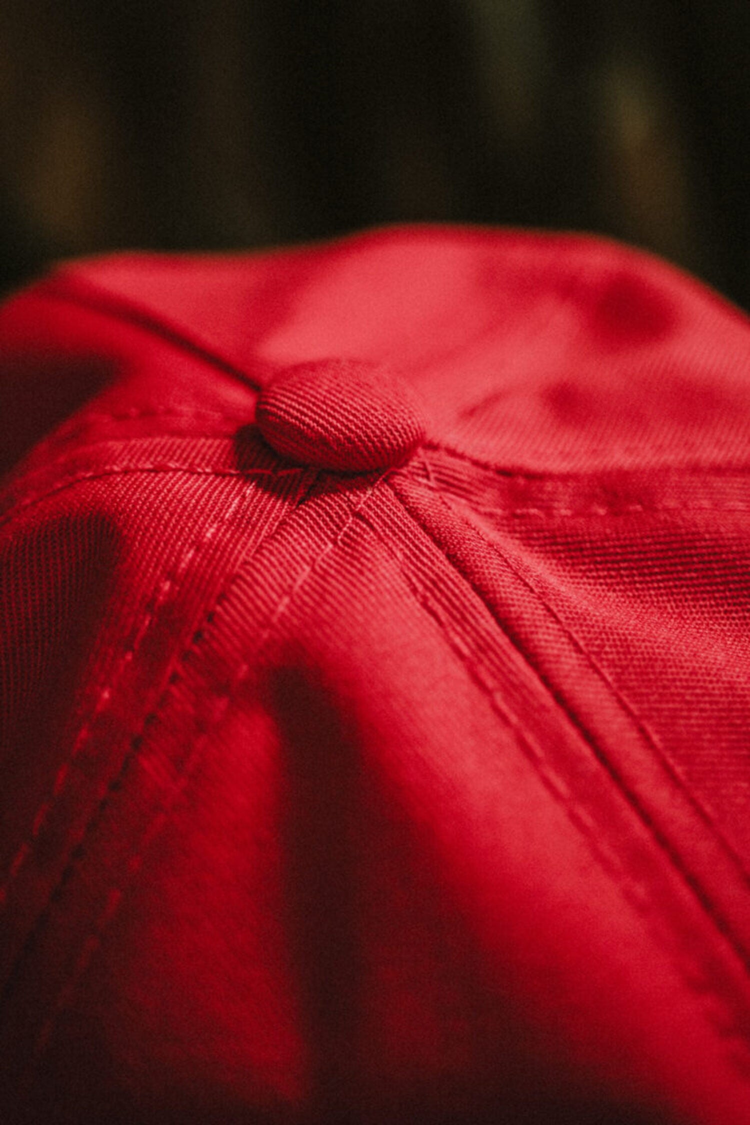 Macro detail of the top of a red cap, focusing on the fabric-covered button and precision reinforced stitching.