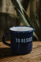 Lobo 'To Resist' navy blue enamel mug, 0.40 L capacity, displayed on a rustic wooden table with plants in the background.
