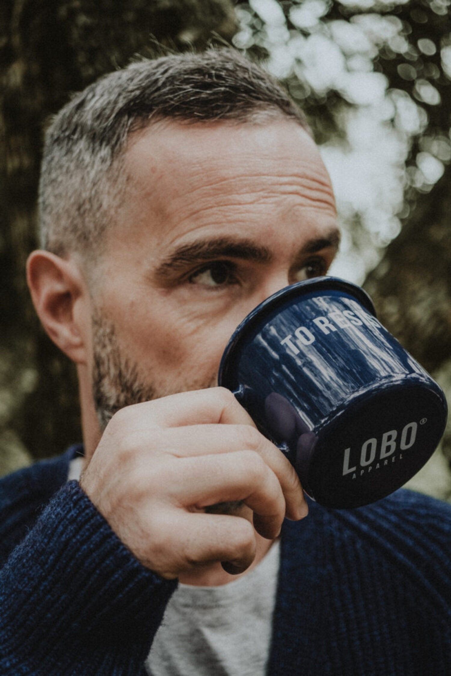 Close-up of a bearded man drinking from the blue 'To Resist' enamel mug, highlighting the Lobo Apparel logo on the base, in a nature setting.