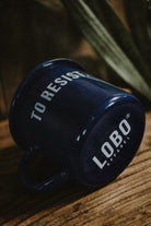 Blue 'To Resist' enamel mug lying on wood, highlighting the Lobo Apparel logo on the base and its glossy finish.