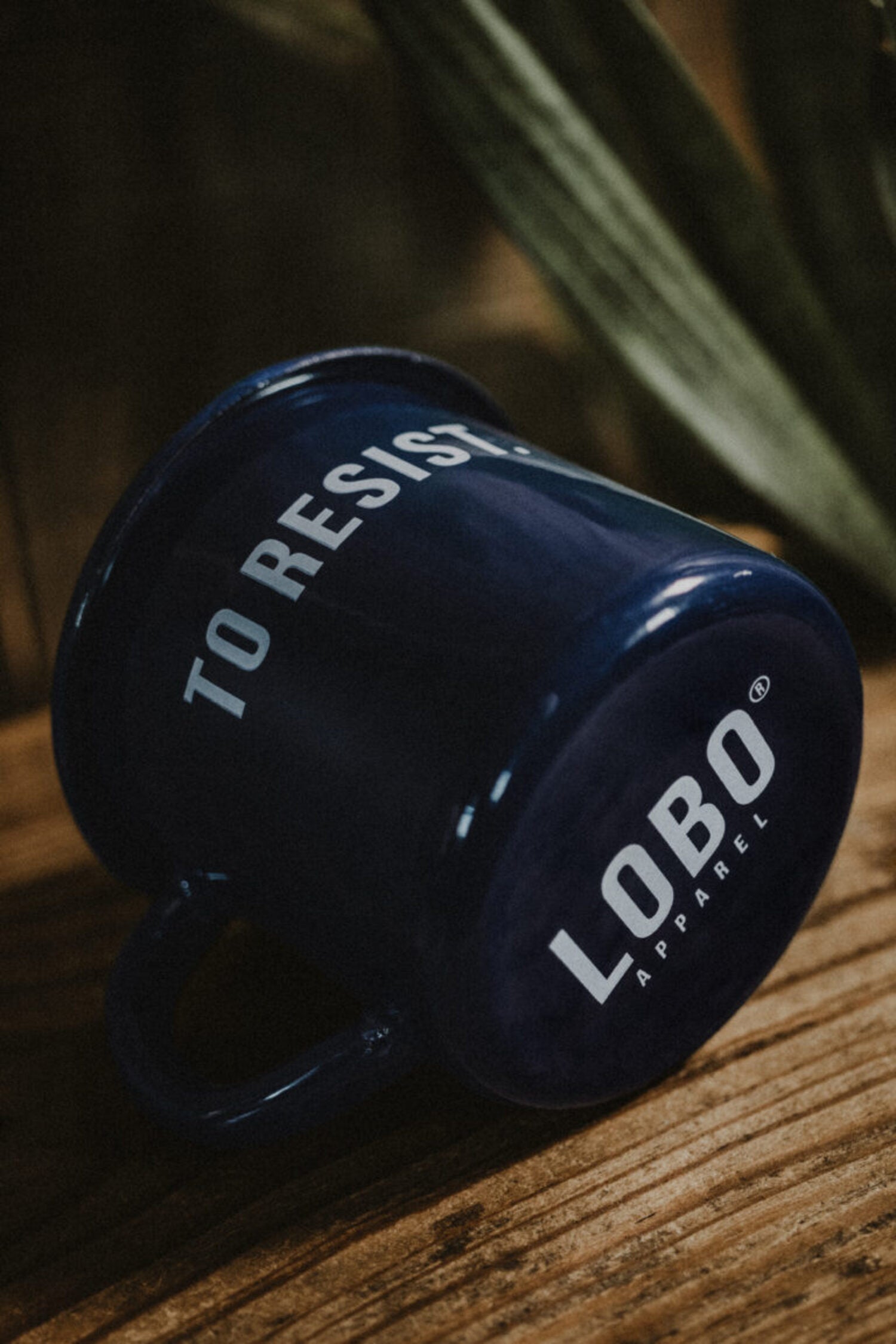 Blue 'To Resist' enamel mug lying on wood, highlighting the Lobo Apparel logo on the base and its glossy finish.