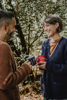 Smiling man and woman in a forest setting, holding Lobo 'To Resist' and 'To Last' enamel mugs, wearing cozy wool knitwear.