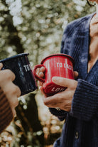 Two people holding Lobo enamel mugs — 'To Resist' in blue and 'To Last' in red — in an outdoor forest setting.