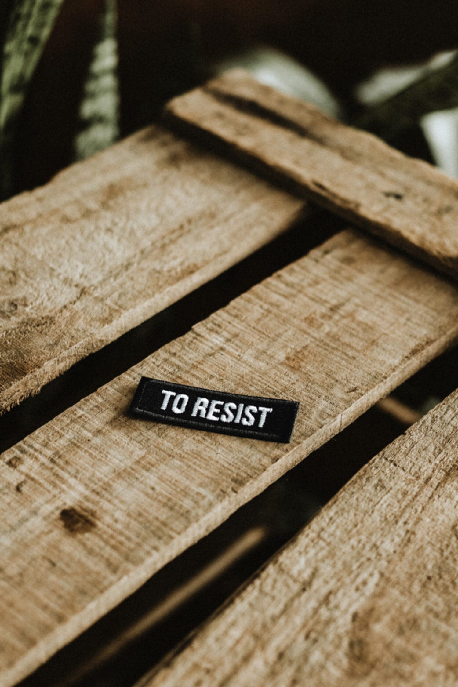 Perspective view of the Lobo "TO RESIST" black embroidered patch on aged wooden slats, durable fashion accessory.