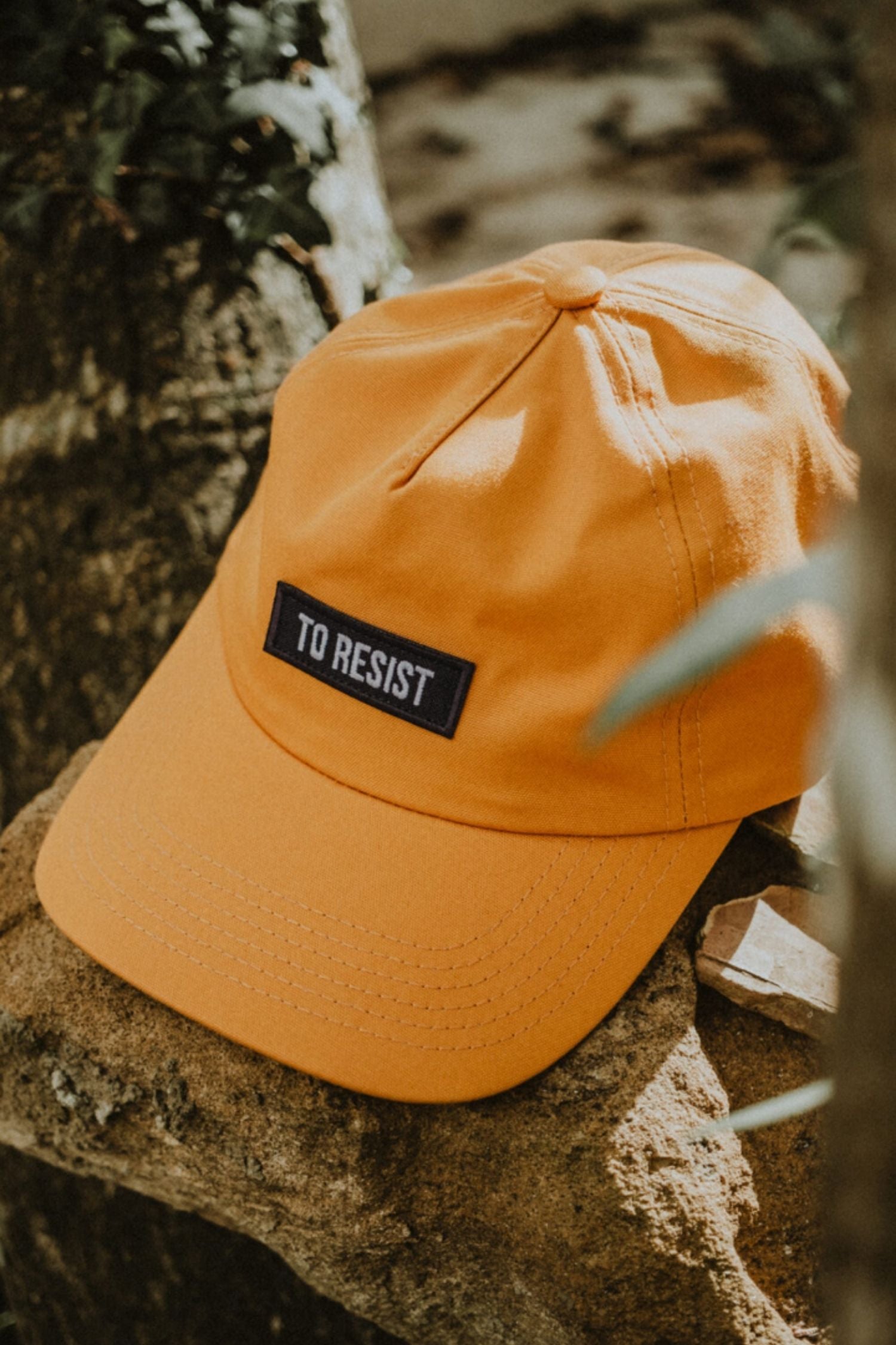 Lobo mustard yellow cap featuring "To Resist" embroidered patch on a natural rock background.