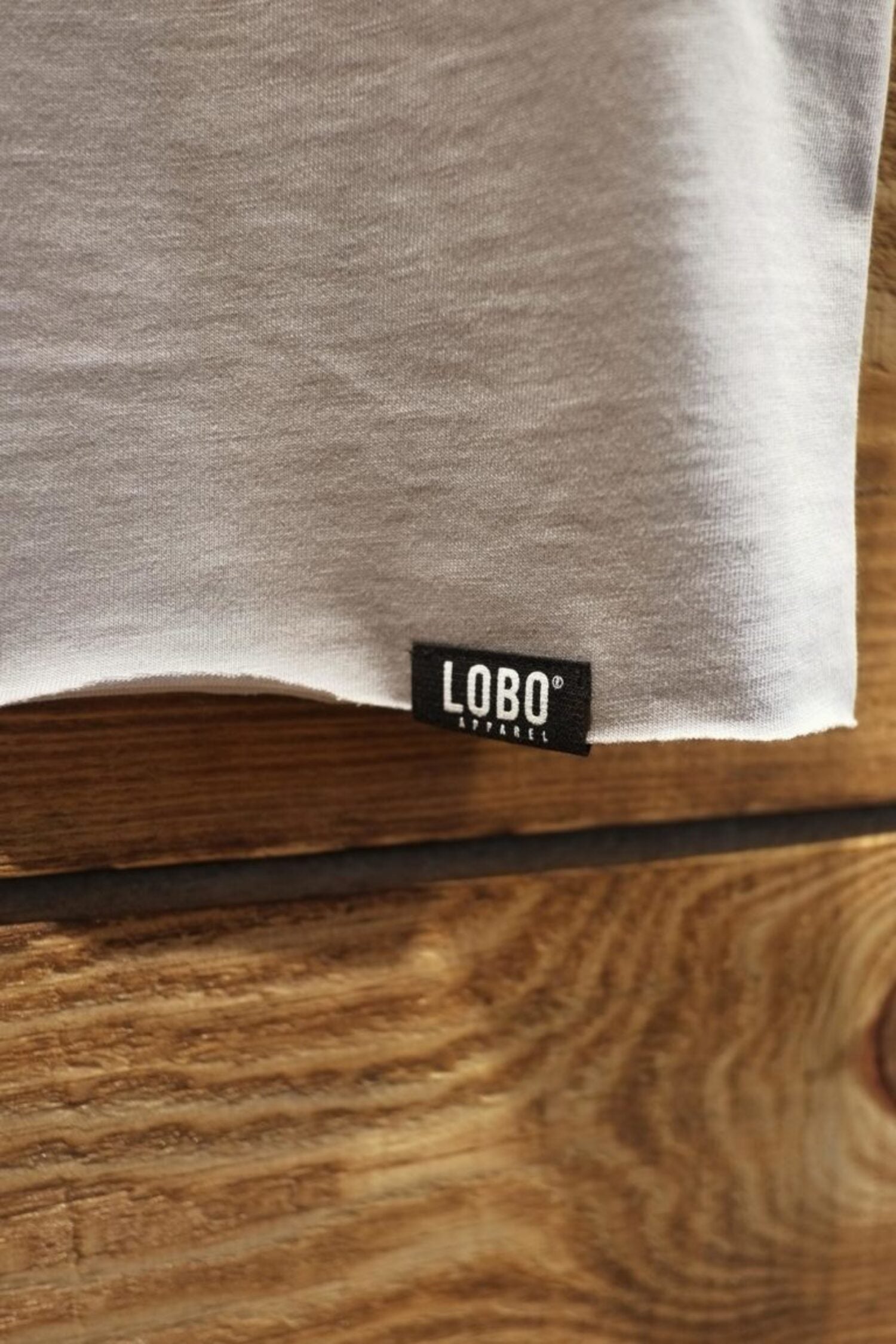 Detail of the black Lobo Apparel woven label and the raw-edge hem finish on the grey t-shirt against a wooden background.